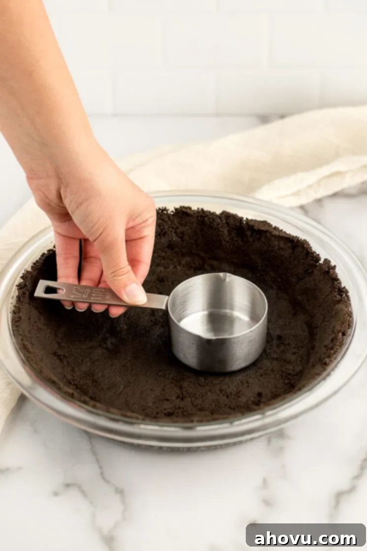 A baker's hands expertly pressing a homemade Oreo cookie crust into a glass pie dish, ensuring an even and firm base.