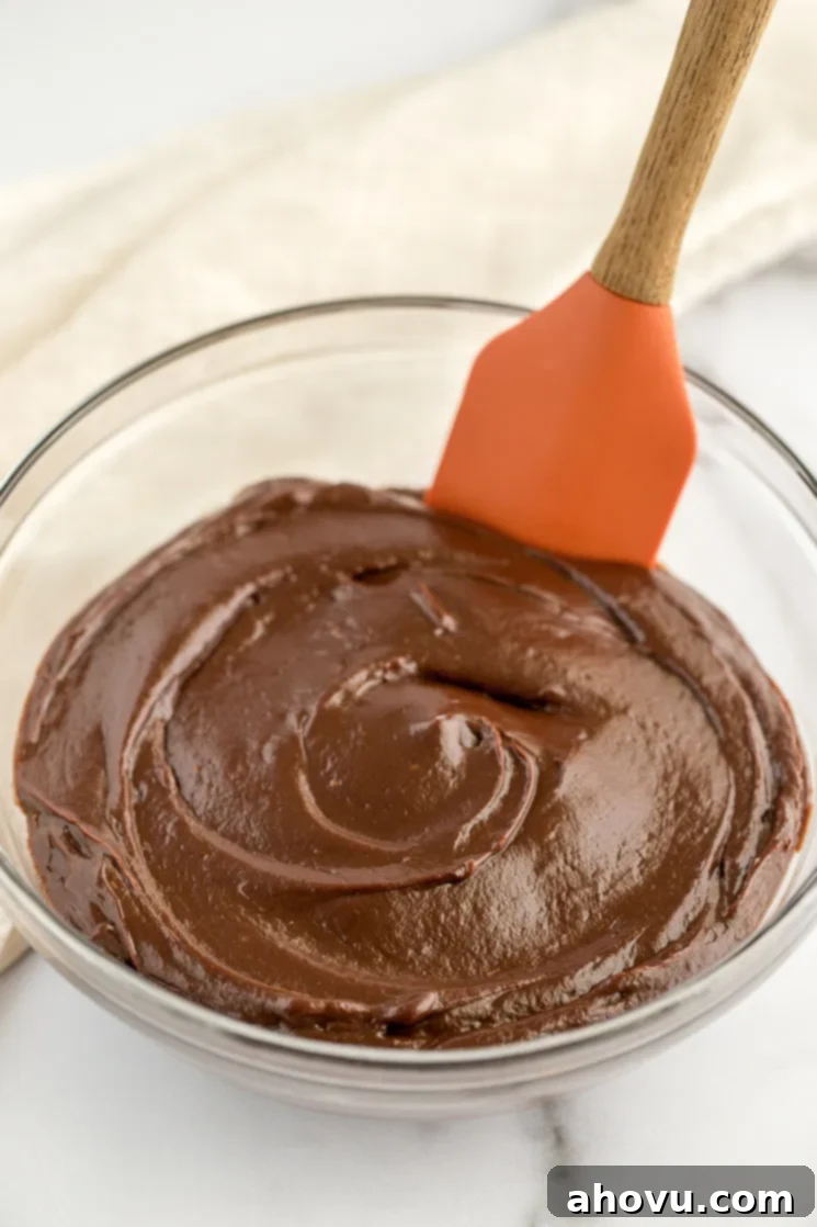 A bowl of glossy, rich chocolate pie filling with a rubber spatula resting on the edge, highlighting its smooth and thick texture.