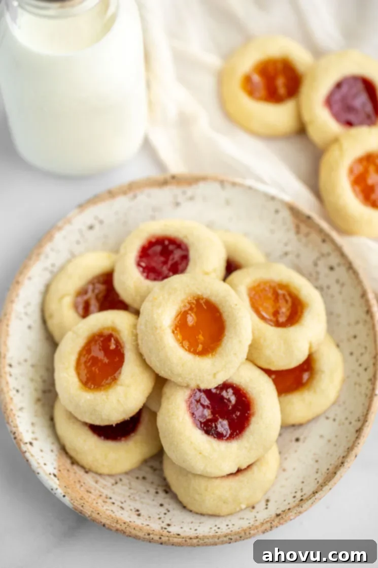 Jammy Jewels 2 A beautifully arranged speckled plate filled with golden-brown thumbprint cookies, featuring both strawberry and apricot jam fillings. More cookies and a glass of milk are visible in the soft-focus background, suggesting a cozy dessert setting.