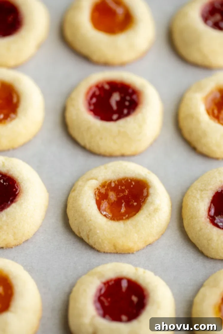 Jammy Jewels 4 A close-up shot of a baking sheet filled with freshly baked thumbprint cookies, perfectly golden-brown with vibrant strawberry and apricot jam centers. The cookies are arranged neatly on parchment paper, highlighting their delicate shortbread texture.