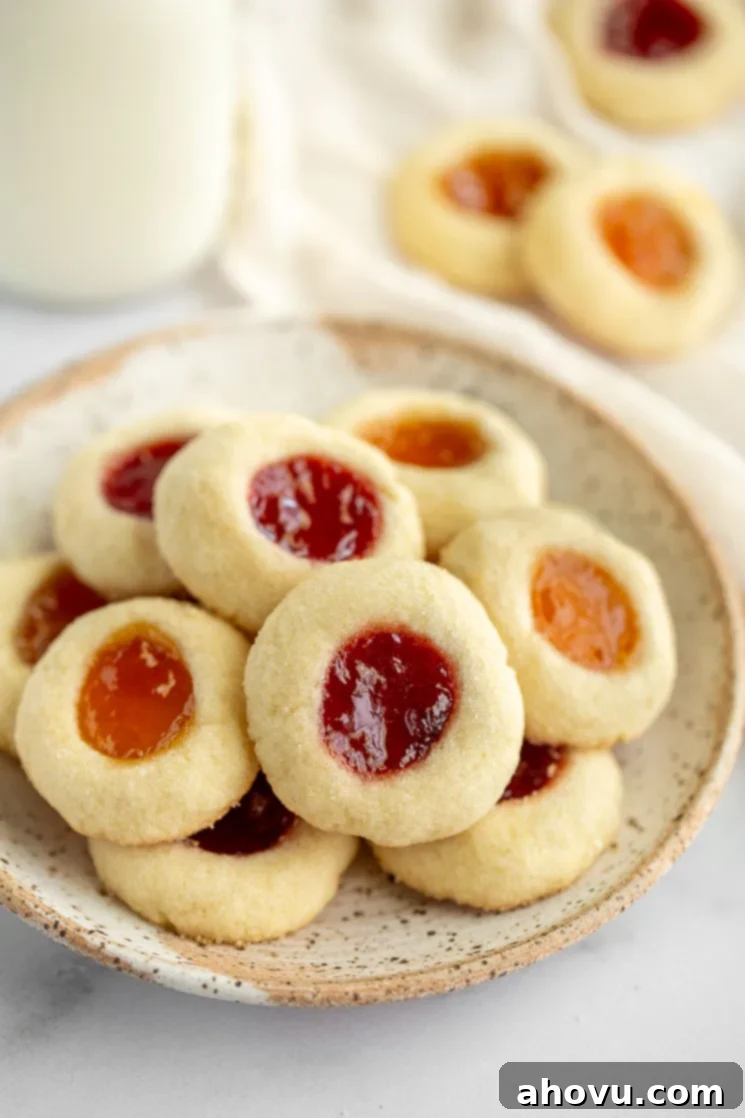 Jammy Jewels 5 A close-up shot of finished thumbprint cookies, featuring vibrant strawberry and apricot jam fillings, arranged on a speckled plate. More cookies are visible in the background, creating an inviting and bountiful display.