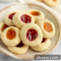 Jammy Jewels 6 A close-up of a single thumbprint cookie, showcasing its buttery shortbread base and a vibrant jam filling, resting on a speckled plate.