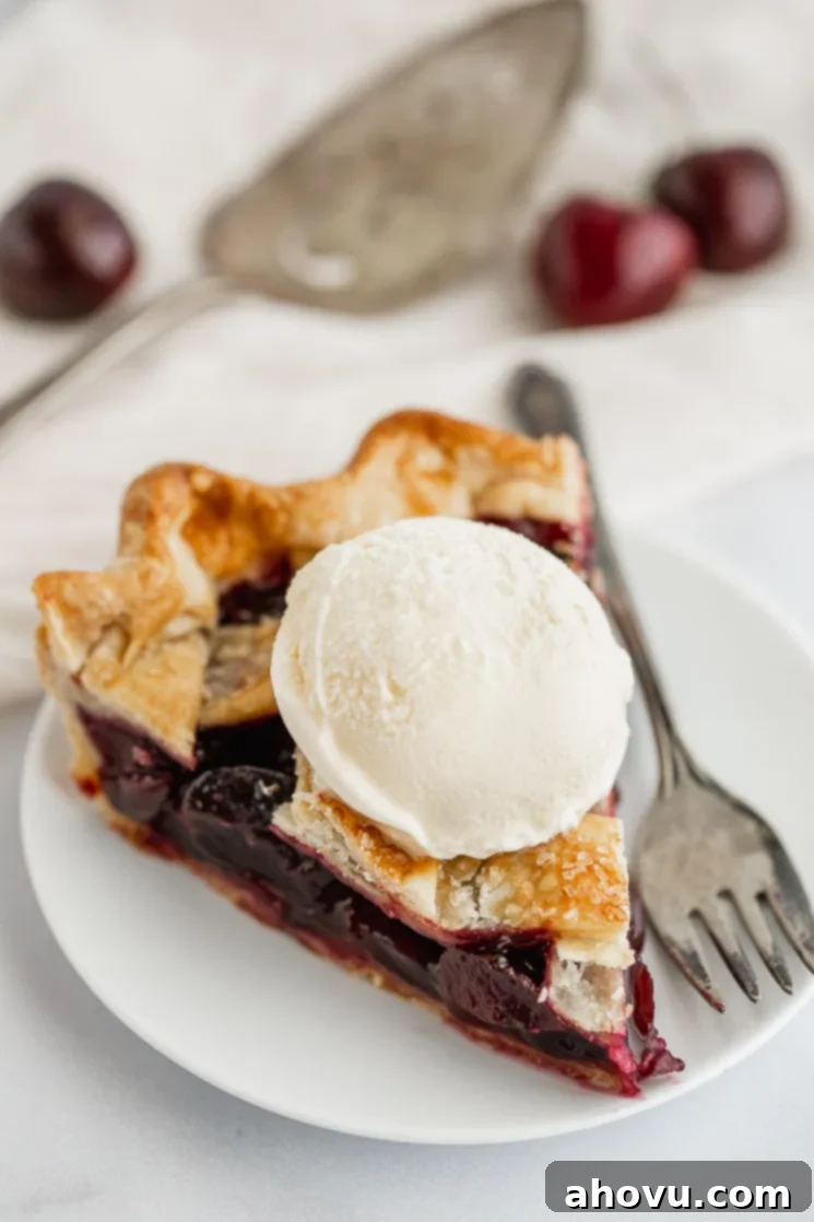 A slice of cherry pie with ice cream on top on a white plate.