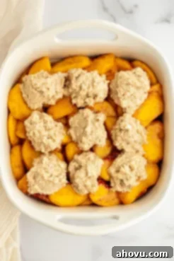 A white baking dish filled with a vibrant peach filling and generously topped with an unbaked biscuit mixture, ready for the oven.