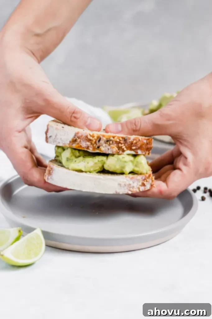 A sandwich filled with homemade avocado chicken salad. Two hands are holding the sandwich up over a gray plate.