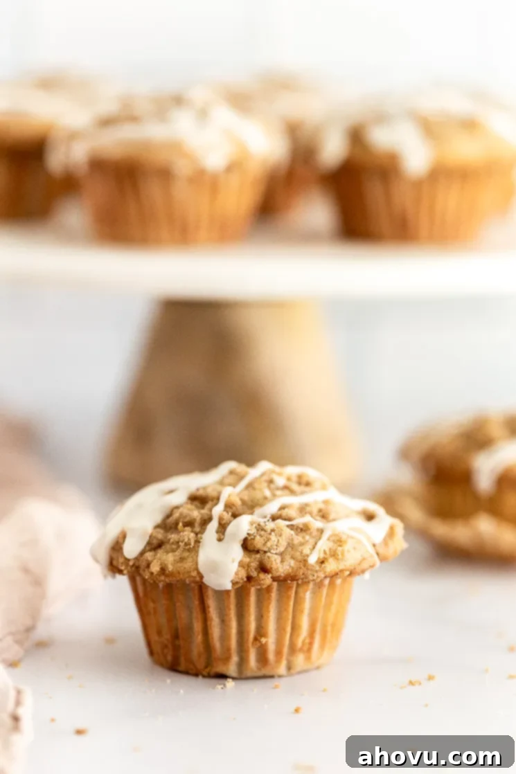 Spiced Apple Muffins 5 A close-up of an apple muffin with a delicate vanilla glaze, showcasing its soft interior and apple chunks. More muffins are blurred in the background on a cake stand.