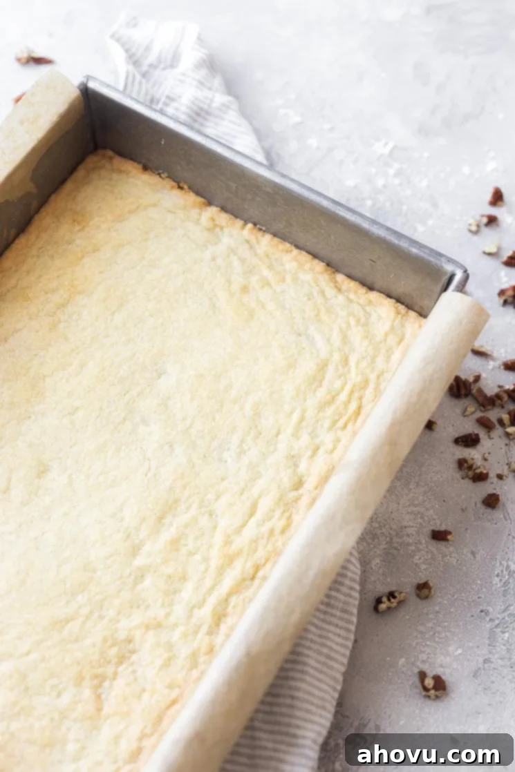 A 9x13 inch baking pan lined with parchment paper, containing a baked, lightly golden shortbread crust, ready for the pecan filling.