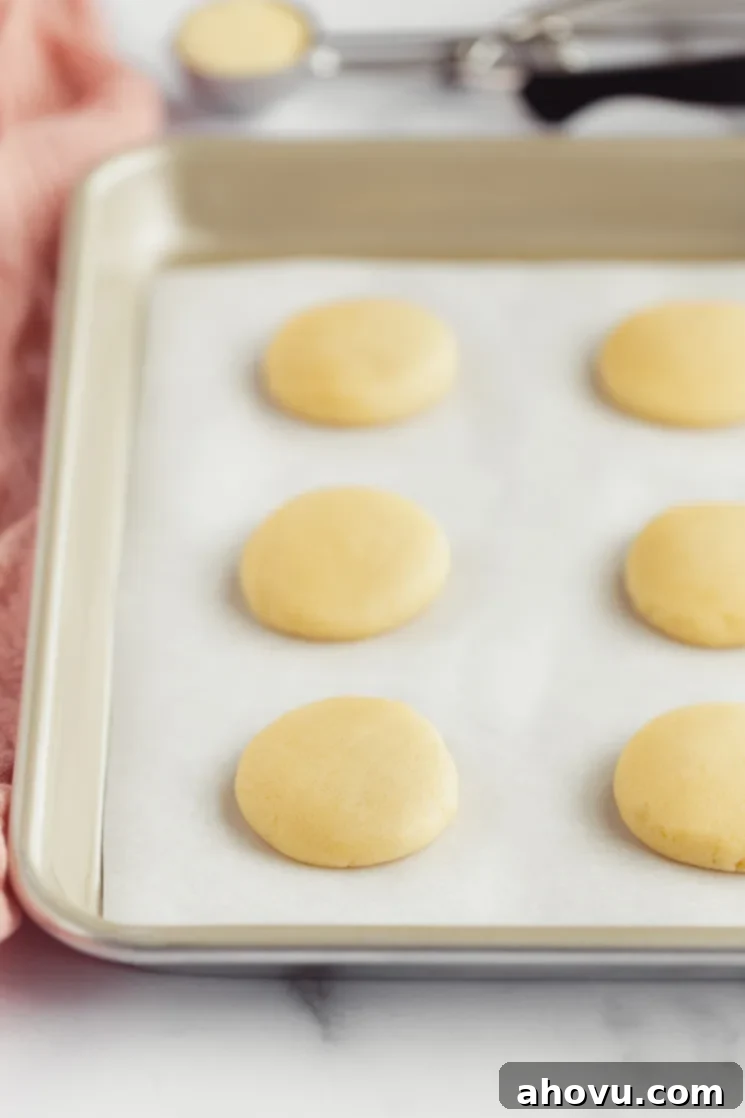 Scooped and gently flattened mounds of easy no-chill sugar cookie dough resting on a parchment-lined metal baking sheet, prepared for baking into soft and chewy treats.