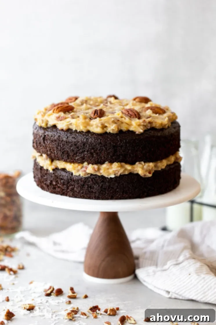 Coconut Pecan Chocolate Dream 2 A beautifully layered German Chocolate Cake on a white marble cake stand, adorned with chopped pecans and shredded coconut around its base. A perfect dessert for any occasion.