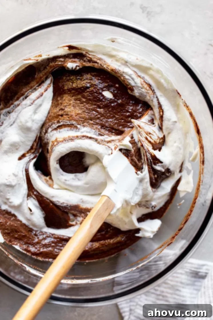 A glass mixing bowl with the chocolate mixture and whipped cream being mixed together with a rubber spatula.