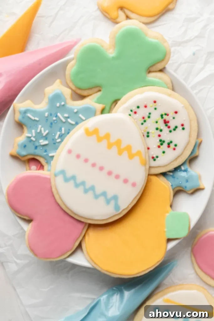 An overhead view of a pile of iced sugar cookies on a white plate. Piping bags of colored sugar cookie frosting are strewn around the plate. 