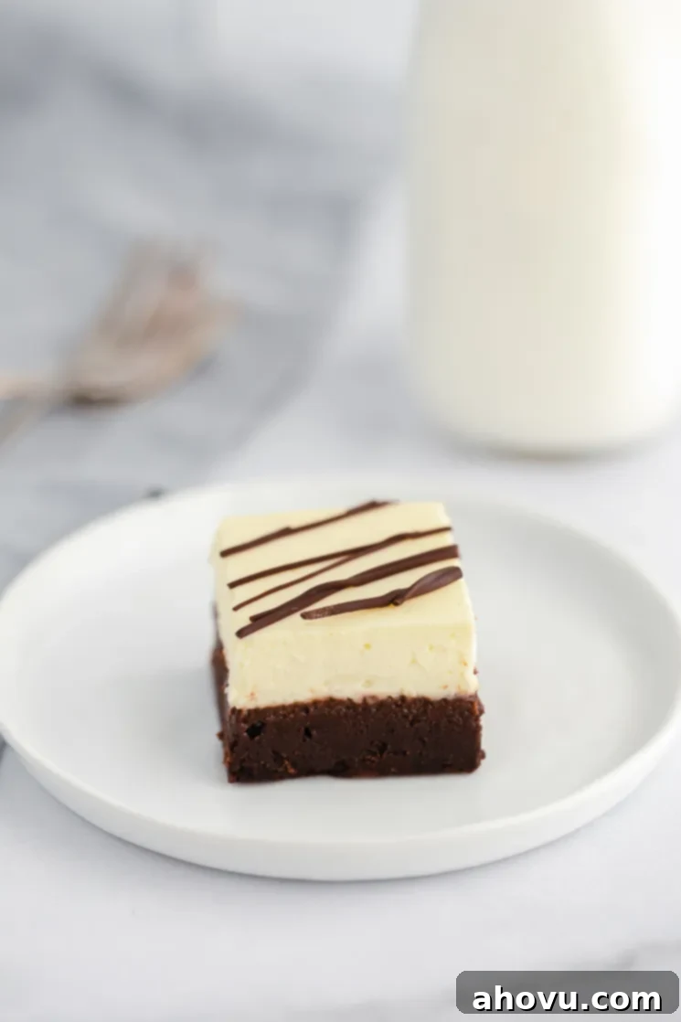 An individual cheesecake brownie drizzled with chocolate on a white plate with forks and milk in the background.
