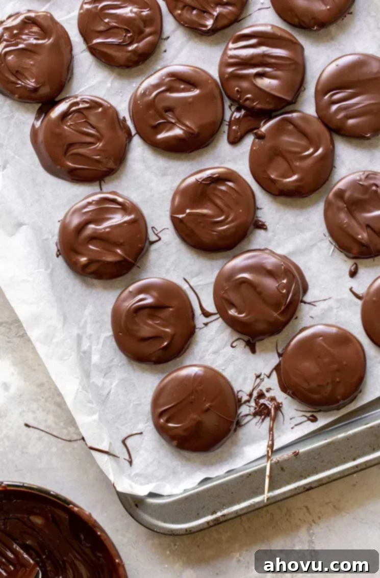 A baking sheet lined with parchment paper, displaying rows of freshly dipped homemade peppermint patties, gleaming with a smooth, hardened chocolate shell, ready for their final chilling in the refrigerator.