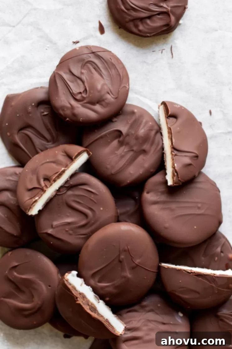 A beautiful display of finished homemade peppermint patties on parchment paper, with several cut in half to showcase the thick, creamy white peppermint filling contrasting with the dark chocolate shell. The overall presentation is elegant and appealing.