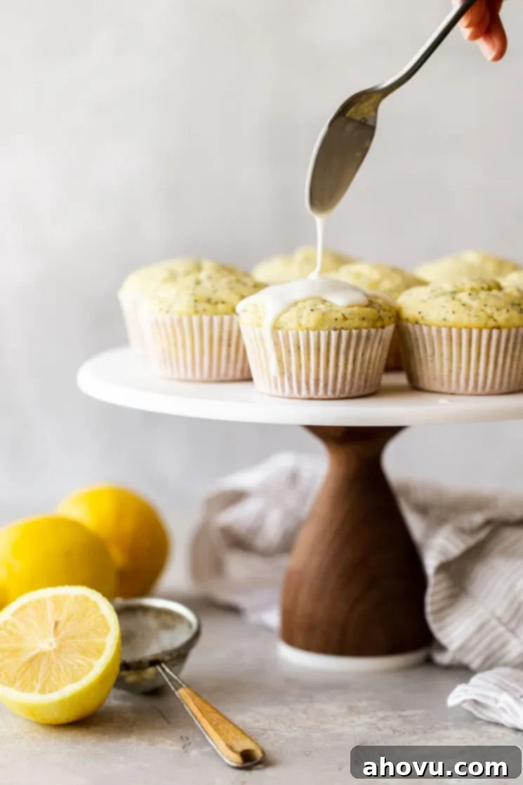 A marble cake stand with lemon poppy seed muffins being topped with an easy lemon glaze.