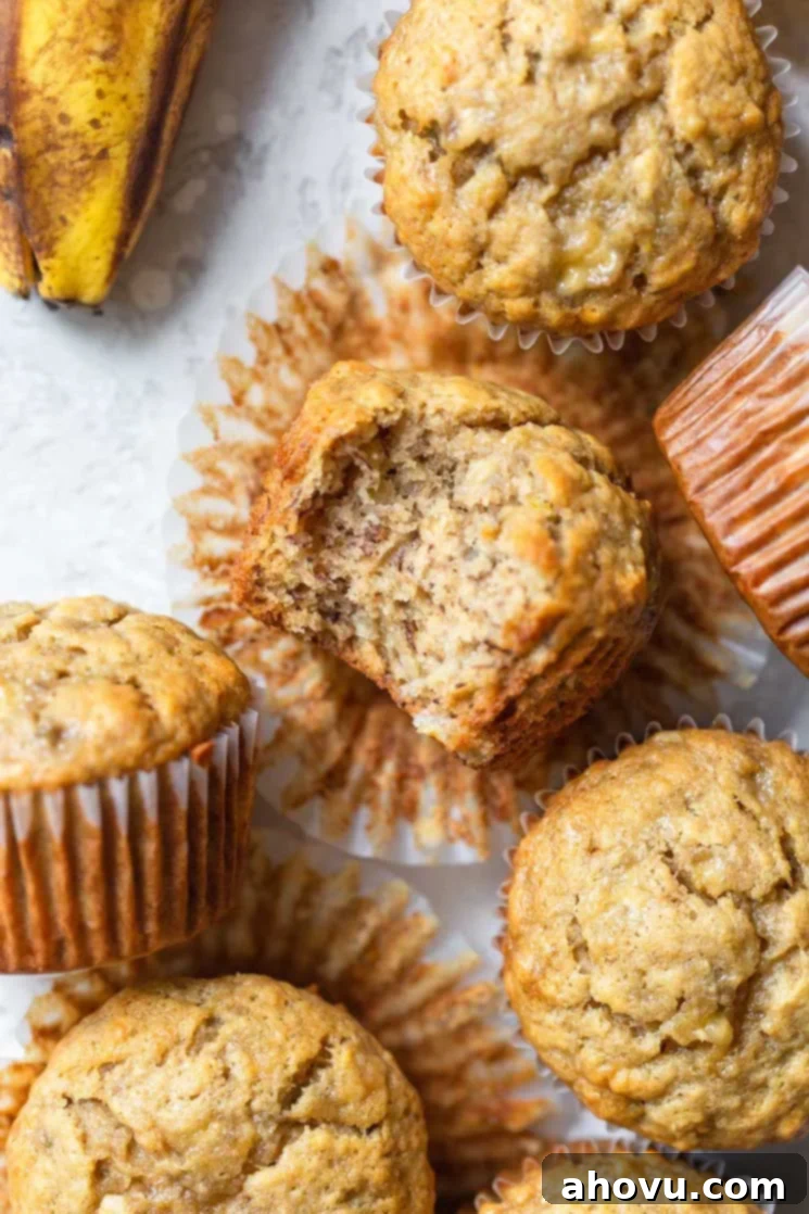 A group of freshly baked, golden-brown banana muffins arranged on a rustic gray surface. One muffin is on its side, revealing its moist, tender interior with a bite taken out, showcasing the perfect texture.