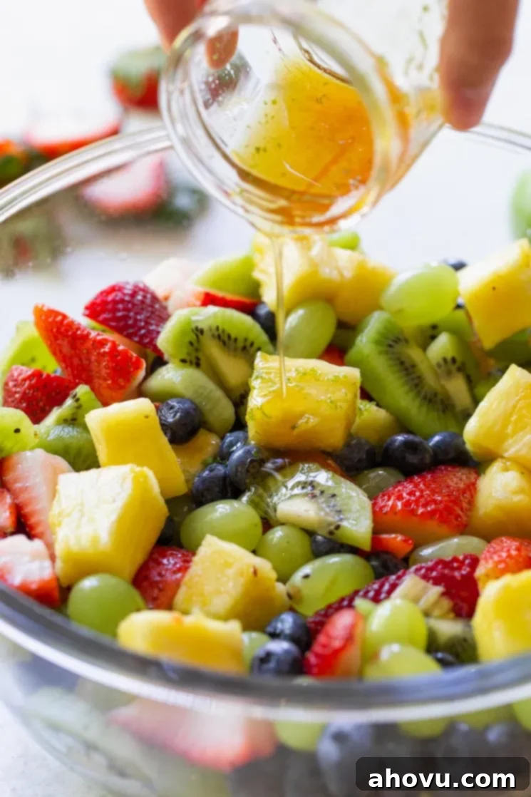 A clear glass bowl brimming with a colorful assortment of fresh fruit salad, while the glistening honey lime dressing is gracefully poured over it from a separate glass jar, ensuring every piece is coated.