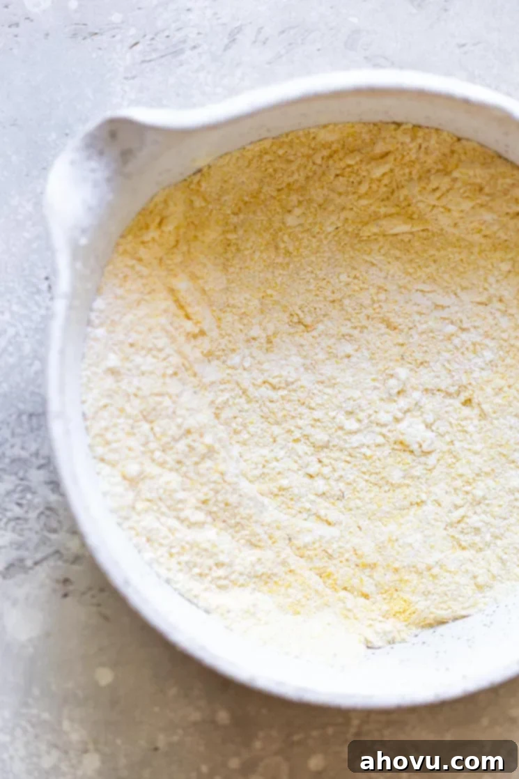 The dry ingredients needed to make cornbread, including flour, cornmeal, baking powder, baking soda, and salt, artfully arranged in a rustic white speckled clay bowl.