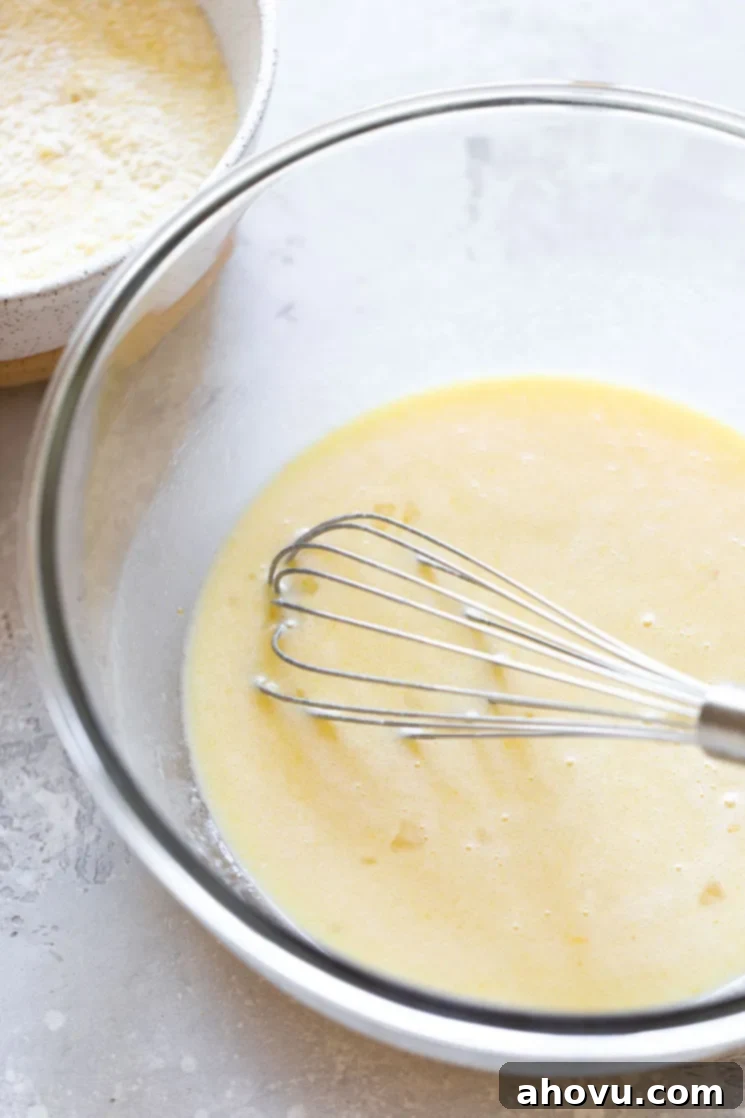 The wet ingredients for cornbread, including melted butter, sugar, honey, eggs, and buttermilk, being mixed in a clear glass bowl, with the rustic bowl of dry ingredients visible in the soft background.