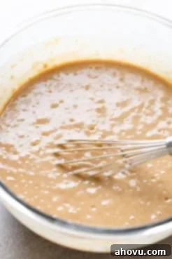 A clear glass mixing bowl contains the rich, brown spice cake batter being thoroughly combined with a metal whisk, showcasing the smooth consistency.