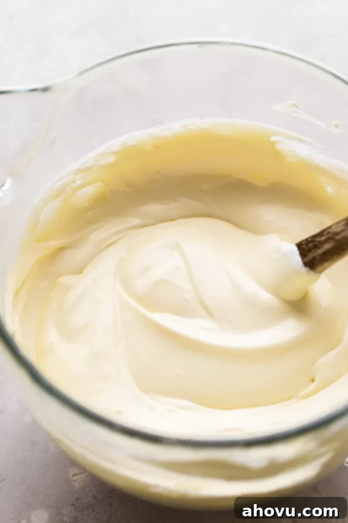 A glass mixing bowl with the finished mascarpone filling after the heavy cream has been folded in. A rubber spatula with a wooden handle rests in the bowl.