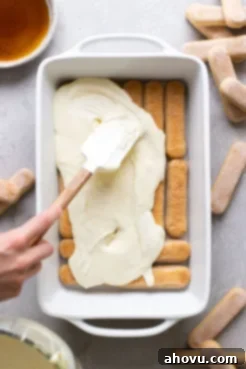 Overhead view showing mascarpone filling being spooned onto coffee-dipped lady fingers in a white baking dish.