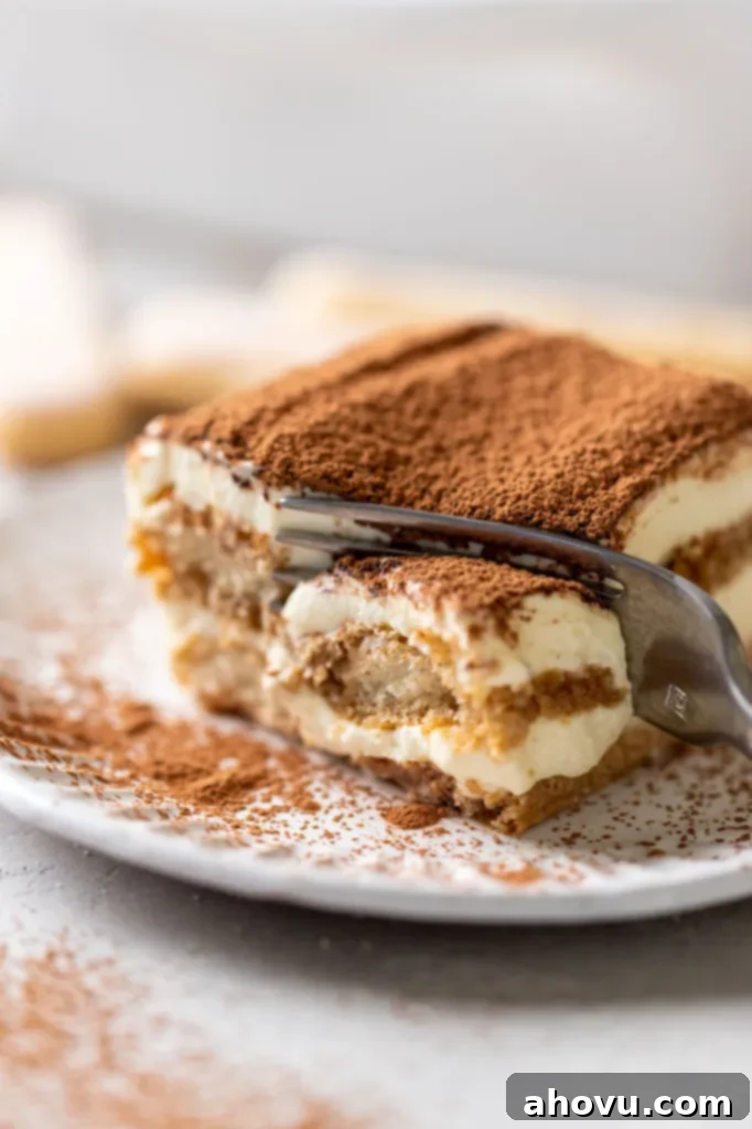 A slice of tiramisu on a white decorative plate. A fork is digging into the end of the slice.