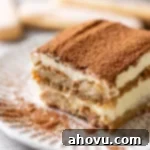 A slice of tiramisu on a white decorative plate. Three lady fingers rest in the background.
