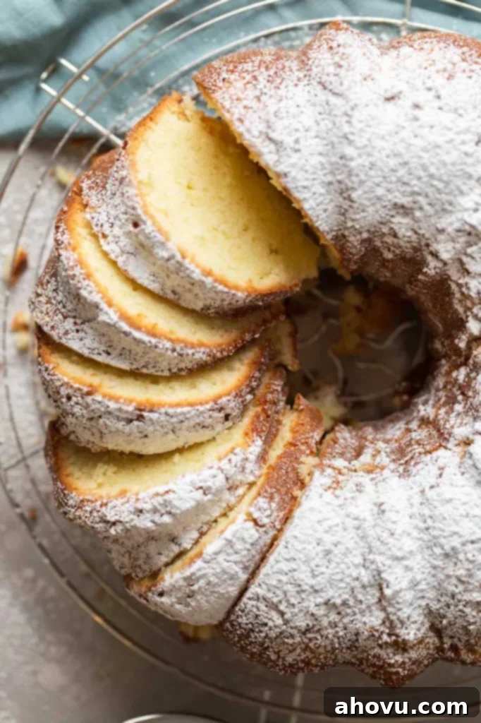 An inviting overhead shot of a freshly baked cream cheese pound cake, lightly dusted with powdered sugar, resting on a wire rack. A portion of the cake has already been elegantly sliced, revealing its moist interior, ready to be enjoyed.