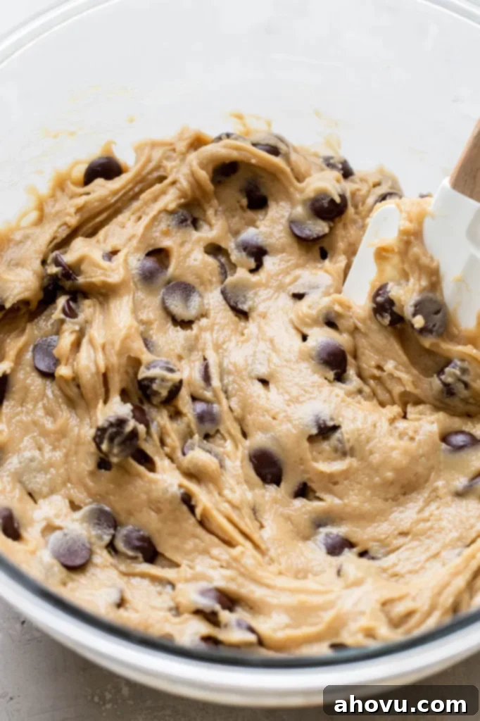 Close-up shot of raw chocolate chip cookie dough in a clear glass mixing bowl, with a red rubber spatula resting in it, ready for baking.
