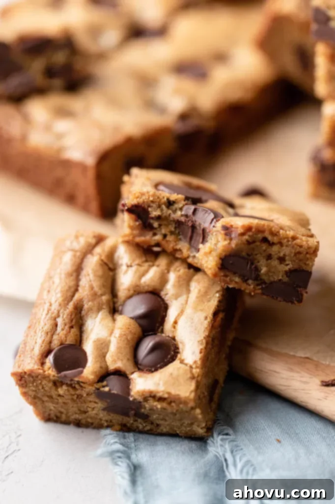 Two perfectly baked chocolate chip cookie bars are resting on each other, with the top bar showing a delicious bite taken out of it. More golden-brown cookie bars are blurred in the background, hinting at a fresh batch.