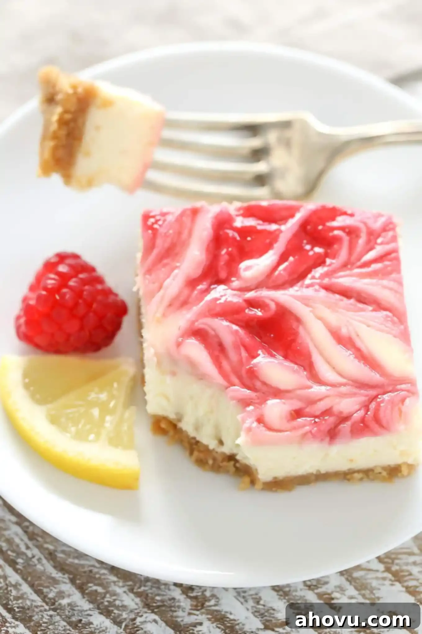 A raspberry swirl cheesecake bar on a white plate. A bite has been speared on a fork, which rests on the plate. There's also a lemon wedge and one fresh raspberry on the plate. 