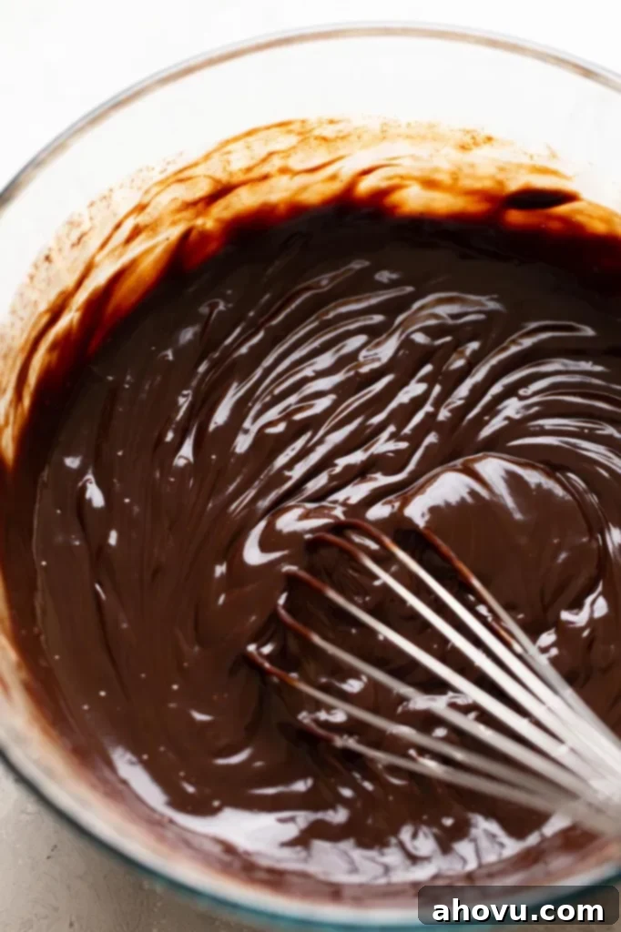 A close-up view of the smooth, glossy chocolate truffle ganache in a clear glass bowl, with a whisk resting inside, illustrating the perfect consistency before chilling.