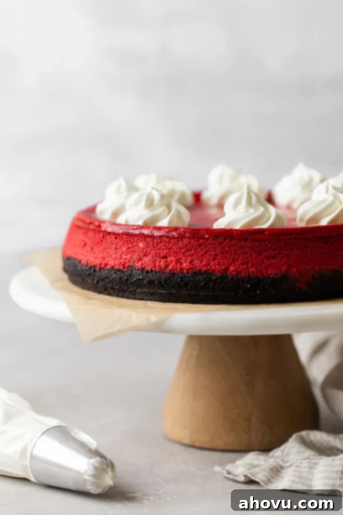 Side view of a red velvet cheesecake topped with whipped cream on a cake stand. A piping bag rests on the side. 