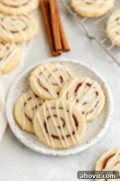 Four cinnamon roll sugar cookies on a speckled white plate. More cookies and three cinnamon sticks rest in the background. Soft sugar cookies with a cinnamon swirl and creamy icing, tasting like miniature cinnamon rolls.