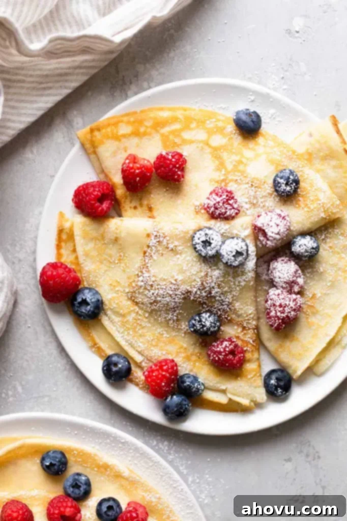 Overhead view of three homemade crepes topped with fresh berries and dusted with powdered sugar on a white plate. Another plate of crepes is partially seen in the lower left corner. 