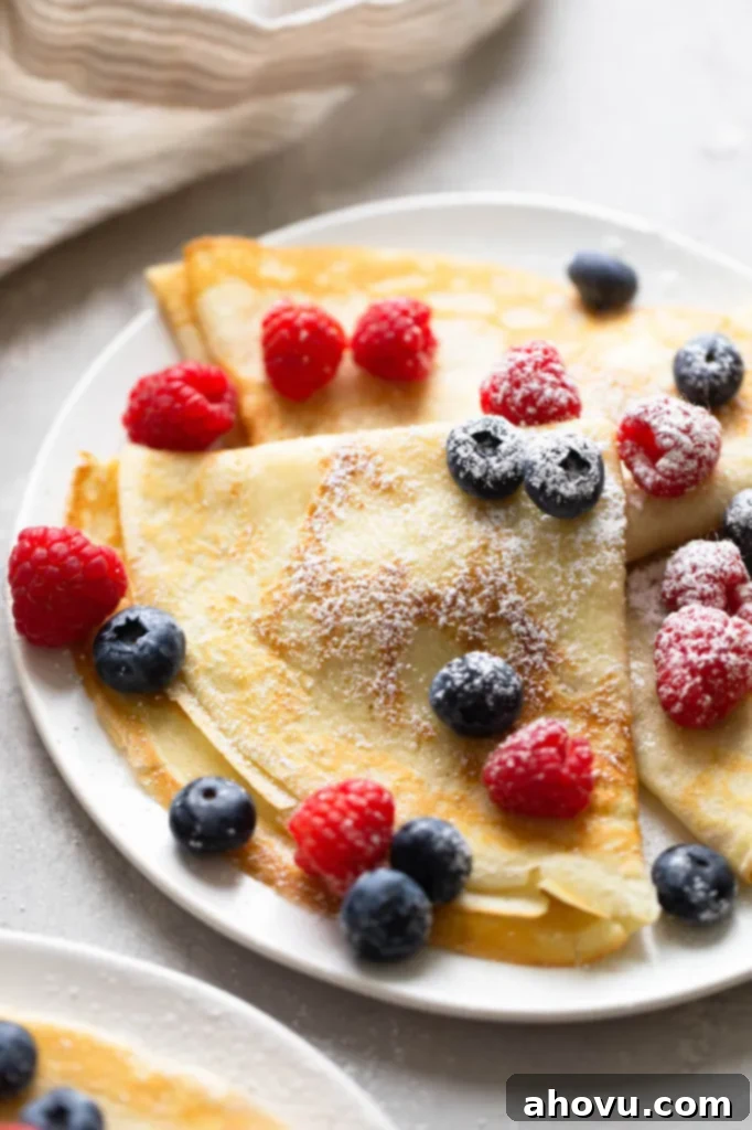 Three crepes topped with berries and dusted with powdered sugar on a white plate. A towel is in the background. 