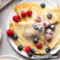 Overhead view of three homemade crepes topped with fresh berries and dusted with powdered sugar on a white plate.