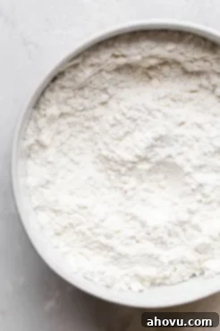 A glass mixing bowl containing all the dry ingredients for muffins: flour, baking powder, and salt. The ingredients are light and well-combined.