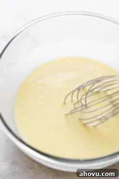 A glass mixing bowl with a whisk, containing the wet ingredients for muffins: granulated sugar, sour cream, oil, lemon juice, lemon zest, eggs, and vanilla extract, all smoothly combined.