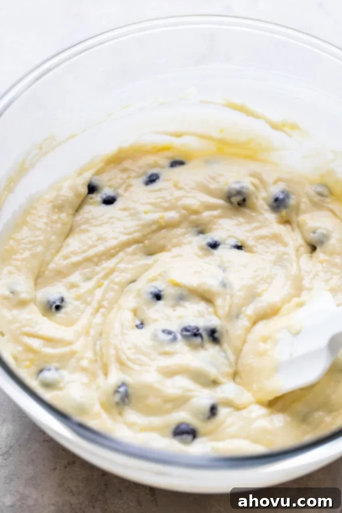 Lemon blueberry muffin batter in a glass mixing bowl. A rubber spatula rests in the bowl, showing the thick, yet airy consistency of the batter with visible lemon zest. 