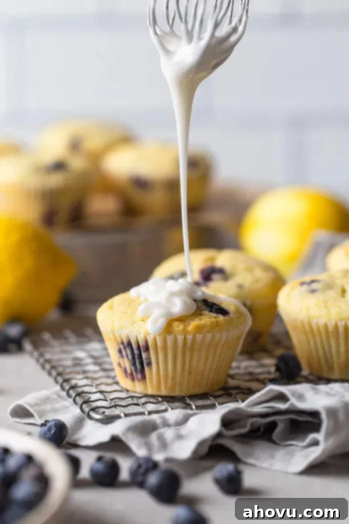A whisk being held over a fully cooled lemon blueberry muffin, with lemon glaze dripping from it. Additional glazed muffins are in the background, alongside fresh lemons and blueberries. 