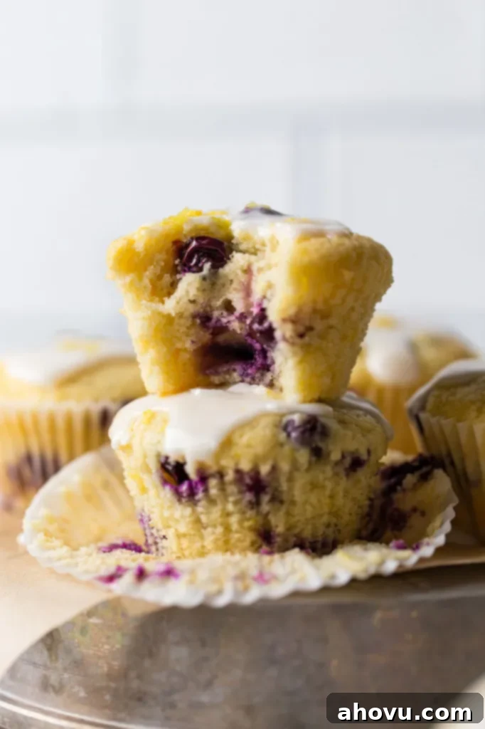 A stack of lemon blueberry muffins on an overturned pie dish, with one muffin having a bite taken out of it, revealing the moist crumb and blueberries. Additional muffins are in the background. 