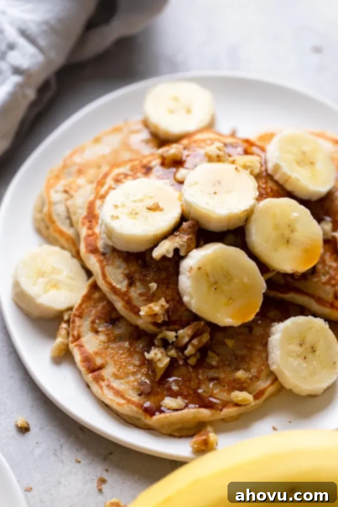 Four easy banana pancakes topped with syrup, banana slices, and walnuts on a white plate. A banana rests in the foreground. 