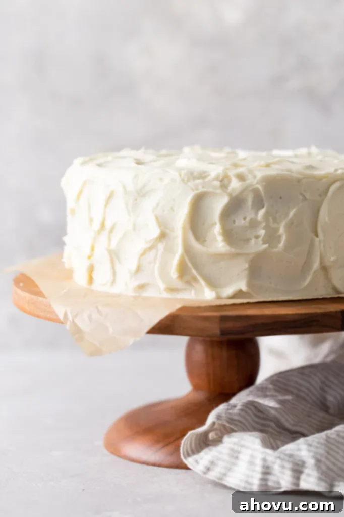 A beautifully frosted homemade vanilla cake sits proudly on a rustic wood cake stand, ready to be sliced. A soft gray tea towel is artfully draped at its base.