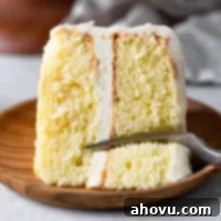 A slice of vanilla cake lying on its side on a wood plate. A fork is digging into the end of the slice.