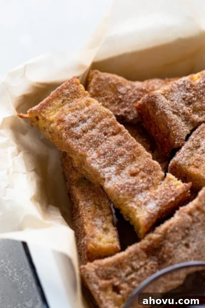 A generous pile of golden-brown cinnamon sugar French toast sticks, fresh from the oven, arranged neatly in a baking dish lined with parchment paper. They look perfectly cooked and ready to serve, inviting a sweet breakfast experience.