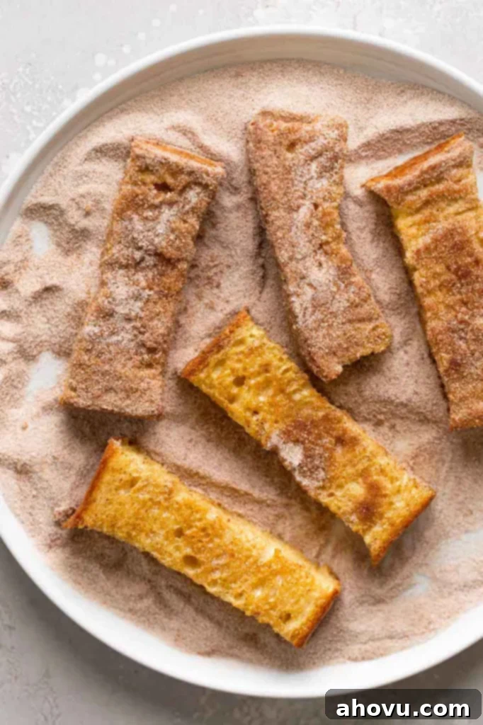 An overhead view of freshly baked French toast sticks being gently rolled in a generous coating of cinnamon sugar on a flat plate, ensuring every side is evenly covered for maximum flavor and visual appeal.