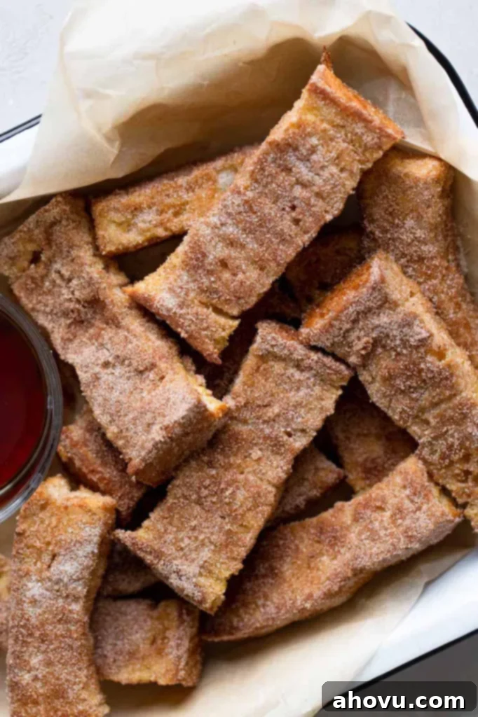 An inviting overhead shot of perfectly baked French toast sticks, generously coated in cinnamon sugar, piled high in a rustic white baking dish. A small, elegant bowl of golden maple syrup is positioned alongside, ready for dipping, promising a delightful breakfast.