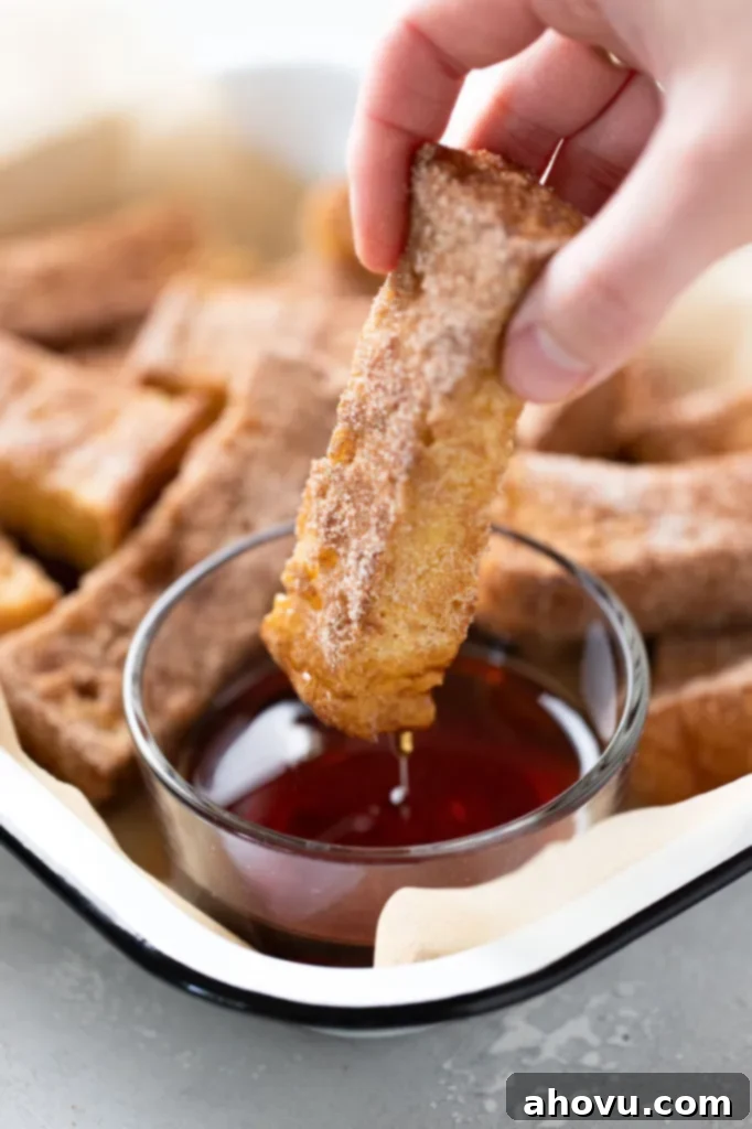 A close-up, mouth-watering shot of a baked cinnamon sugar French toast stick being deliciously dunked into a small, clear dish of rich, golden maple syrup, highlighting its perfectly crispy coating and the tempting liquid sweetness.